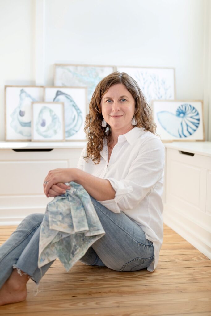 Meet Laura, a local watercolor artist sitting on a wooden floor with her coastal artwork displayed in the background showcasing oysters, shell, and marine plant life in hues of blue