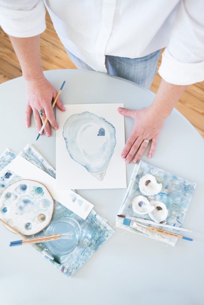 artist holds a paintbrush and uses hands to frame her watercolor painting of an oyster using hues of blue in north carolina