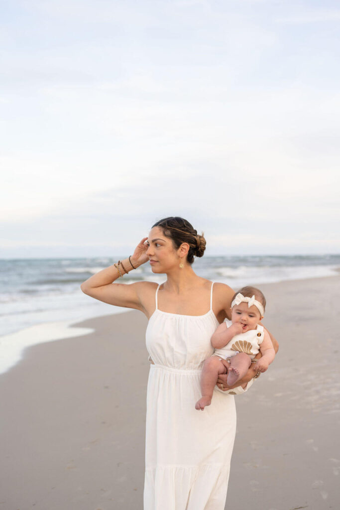 Mom enjoys a quiet walk on the beach with her baby daughter in neutral linen outfits.