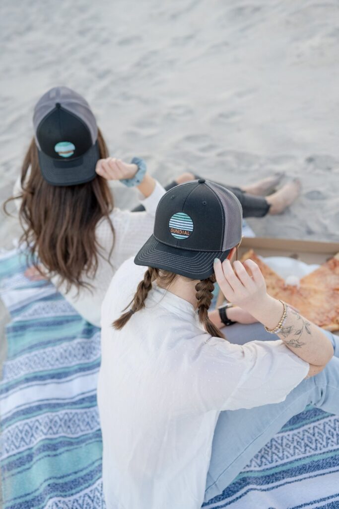 Meet Annie, a content queen with drive and spunk sitting on the beach with a friend wearing backwards baseball caps in Wilmington, NC