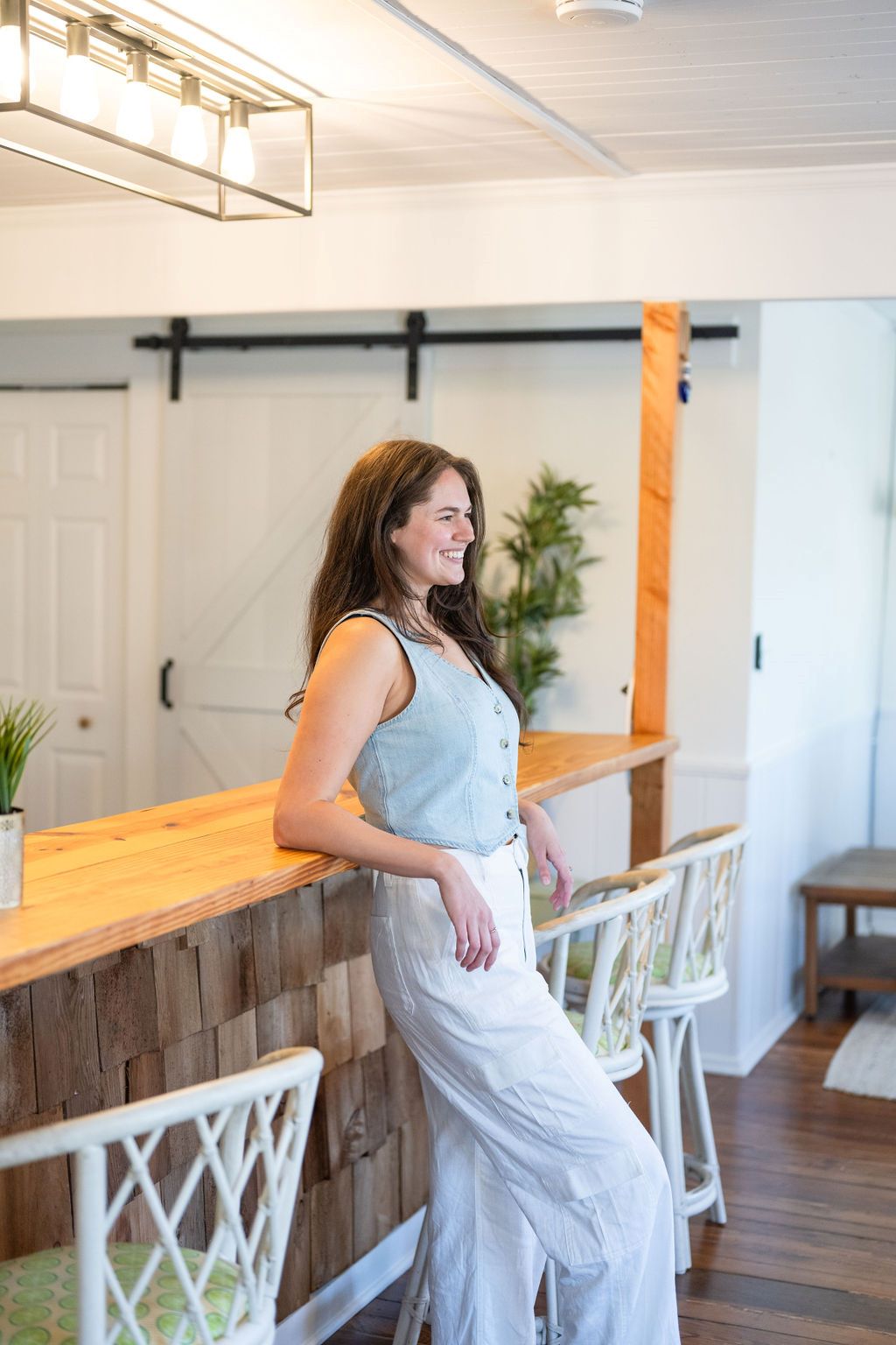 Annie leans up against a wooden bar with her elbows up wearing a denim tank in Wilmington, North Carolina