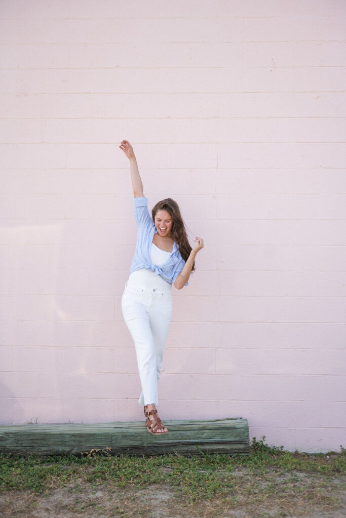 Annie posing with one arm up wearing a denim shirt and white pants in front of pink wall in Surf City, North Carolina